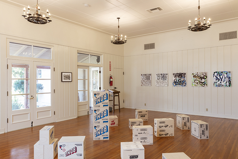 the interior of the art in the park space, screen-printed wood soap-boxes stacked on the floor, and photographs of signage marked with graffiti. three small chandeliers hang from the ceiling