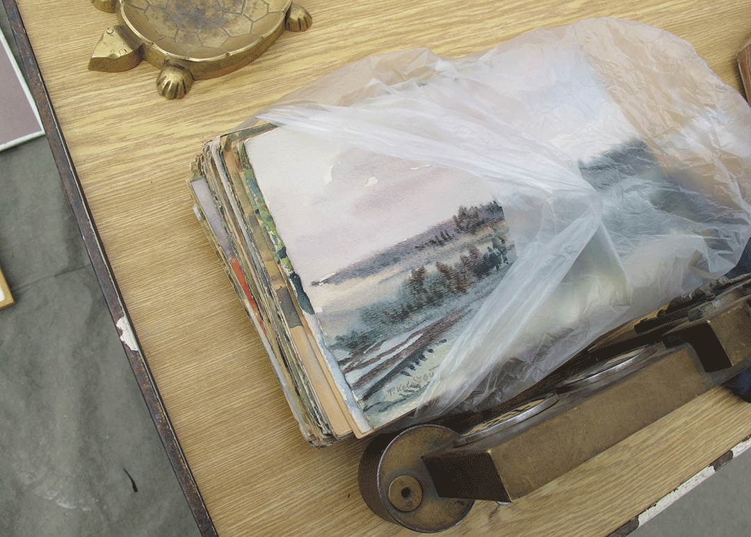 a pile of small watercolor paintings in a clear plastic bag, sitting on a wood surface, next to a gold metallic turtle paperweight / ashtray