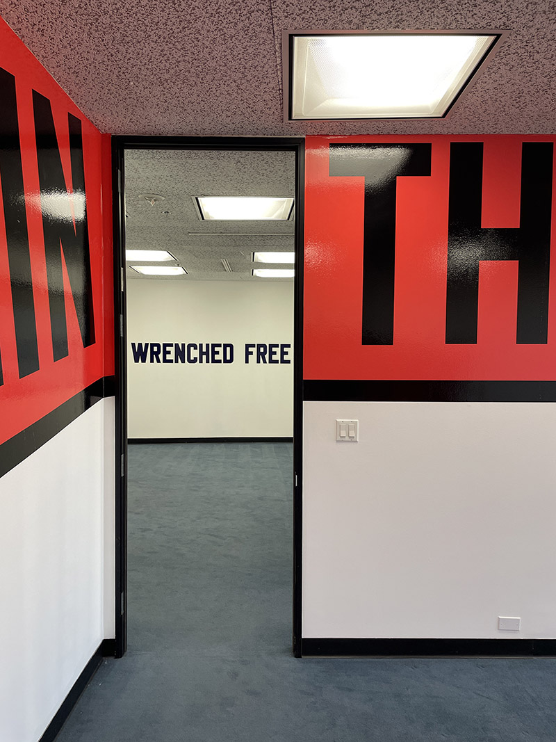 Exhibition view of Lawrence Weiner Wrenched Free at Manual Arts, looking through a doorway, large black letters visible that read IN on the left and TH on the right, with the words WRENCHED FREE visible on a wall through the doorway across another room