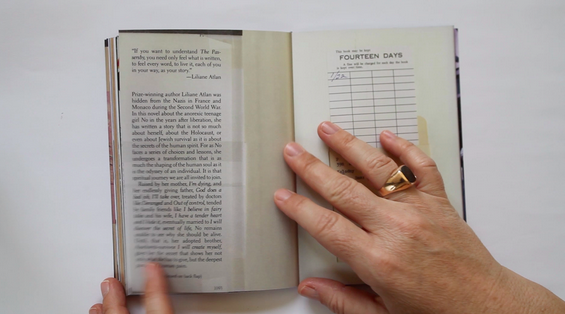 two hands holding open a small library book, left page has a blurb on it, right page has a due-date card. The right hand has a large gold ring on the right ring finger